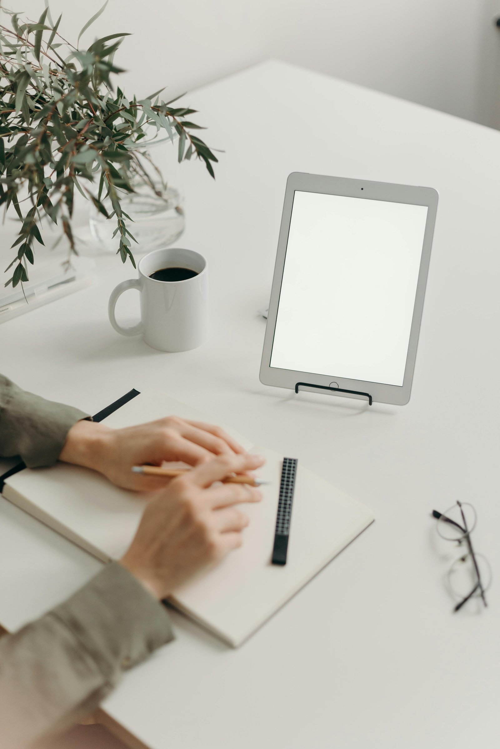 Person writing in a notebook next to a tablet, coffee, and plant